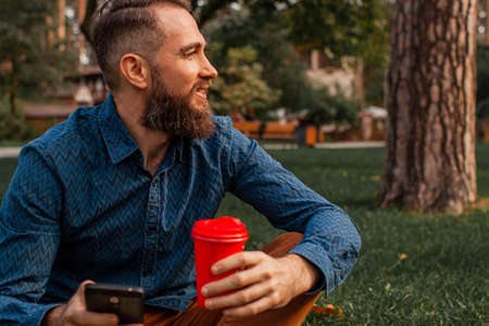 handsome middle-aged bearded man with paper cup of morning coffee walking in autumn park.の写真素材