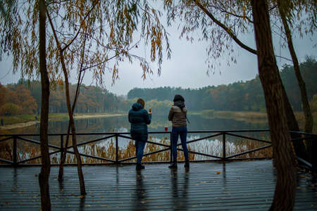 Two female friends in warm coat standing near lake with autumn forest and lake on the background. Cozy fall atmosphere.の写真素材