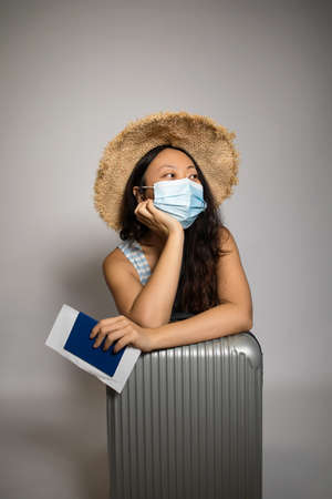 Asian female tourist in medical mask with suitcase on white background. Traveling during coronavirus pandemicの写真素材