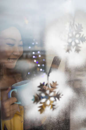 Asian woman looking out the window drinking coffee and using phone in Christmas holidays.