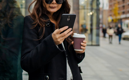 Charming young asian woman waiting for a phone message while drinking coffee in the street. Stylish girl in coat posing in the city, holding a smartphone and enjoying a cappuccino.の写真素材