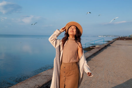Young happy asian woman with hands in the air walks on the seaside in autumn. Seagulls flying on the...