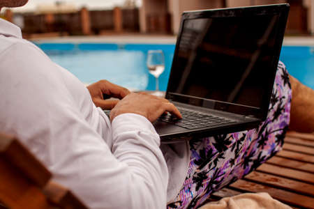 Man lying in chaise-lounge by swimming pool and checking e-mails on laptop. Positive man using laptop on summer vacation.の写真素材