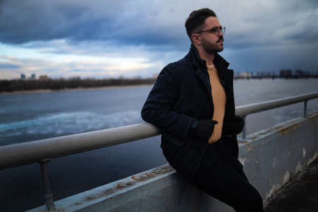 A bearded man with glasses in a winter coat, standing leaning on the railing of a bridge over a...