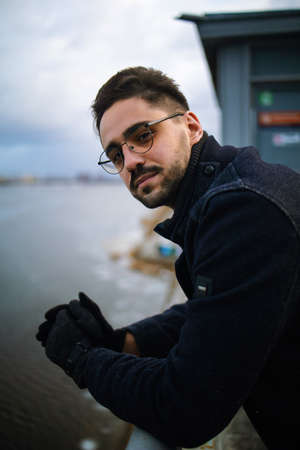 A bearded man with glasses in a winter coat, standing leaning on the railing of a bridge over a canal, deep in thought, looking at camera.の写真素材