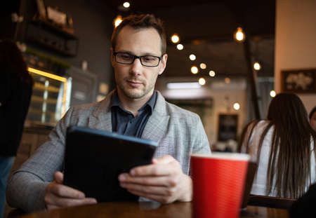 Closeup of serious businessman wearing suit and using tablet in coffee shop, successful manager...