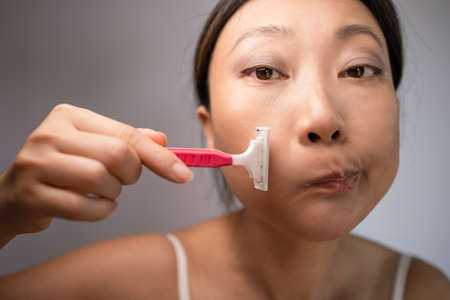 Funny asian woman shaving her face by razor on white background. Pretty woman shaving razor on her...