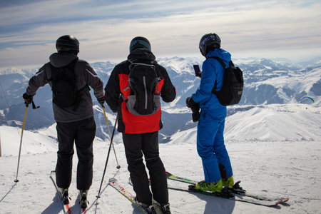 Three hikers with backpacks are on a high snow-covered peak above the clouds in the mountains. They rest after a heavy ascent, enjoying an incredible view.の写真素材