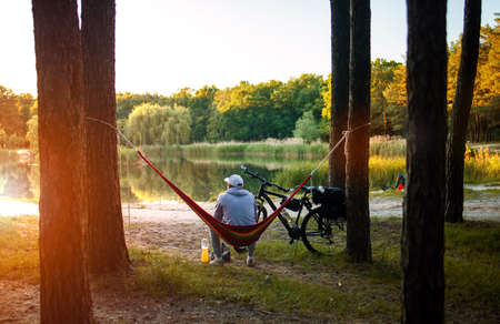 Man travels on bicycle, relaxing in red hammock, rest in forest near lake. Cyclist in hammock at campsite by river. Male on bike in hammock at sunset.の写真素材