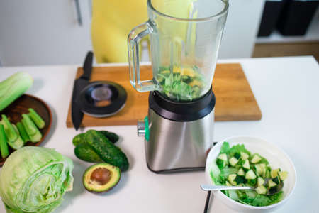 Fit girl preparing green detox juice with spinach and celery at home after training, wearing sport...