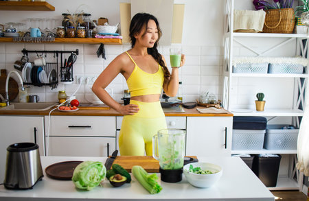 Fit girl preparing green detox juice with spinach and celery at home after training, wearing sport clothesの写真素材