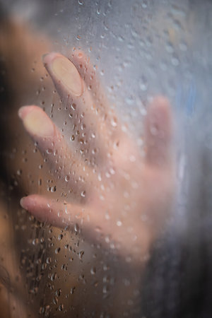 Female hand and unrecognizable silhouette behind wet glass in showerの写真素材