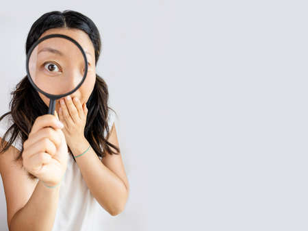 Funny image of young surprised asian woman looking at camera through magnifying glass, isolated on white backgroundの写真素材