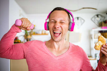 Funny grimacing man working out with small pink dumbbells at home eatting donut