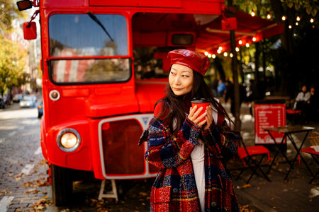 beautiful asian woman in long plaid coat and red leather hat looking with smile while standing near red bus. Street photo of a beautiful woman drinking coffee in front of a cafe anの写真素材