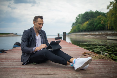 Cheerful and successful businessman works outdoors on wooden bridge near lake holds a tablet