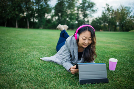 Young asian woman with digital tablet sits on the grass in the park. she using tablet online...