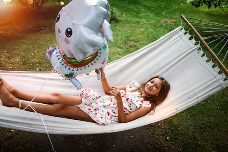 portrait of a little cute girl with long hair resting on a hammock in the garden and smiling.の写真素材