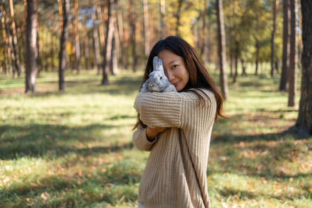 Adorable young rabbit and woman sit together outdoors in autumn forest. Asian woman wear warm sweater with pet in relax and calm.の写真素材