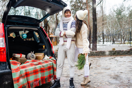 Young beautiful couple in beige sweater standing near car with Christmas decor in winter forest.の写真素材