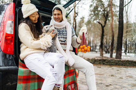 happy couple with rabbit outdoor. Happy couple holding a fluffy rabbit in the back of car with Christmas gift box in winter forest.の写真素材