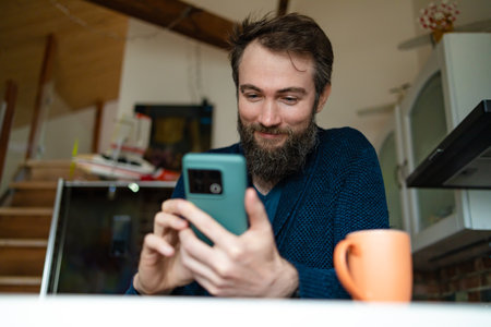 Shot of a handsome young man drinking coffee at home. Happy man drinking a cup of coffee at home. Shot of a young man using a smartphone and having coffee in the kitchen at home.の写真素材