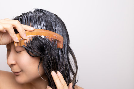 Smiling young Asian woman applying hair mask in a bathroom; haircare concept.