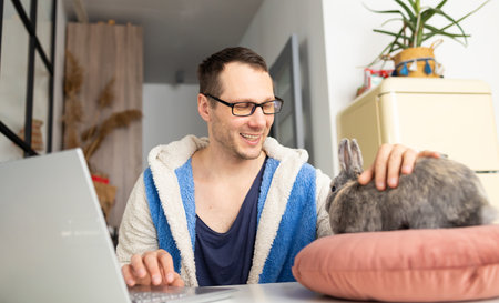 Freelancer man with his friendly bunny using laptop at remote home office. owner and rabbit together. pet adoption.の写真素材