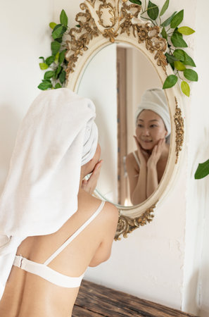 Happy confident young asian lady with towel on head looking at bathroom mirror, touching moisturized soft healthy sensitive skin, doing morning hydration, spa beauty treatment, facの写真素材