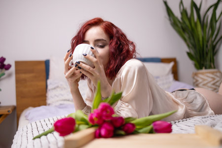 Young red head girl holding a mug of hot coffee in hands. spring breakfast with tulips on bed.の写真素材