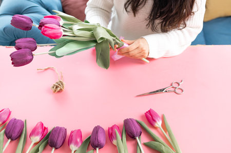 Female's hands wrapping a bunch of pink tulips next to scissors and jute rope on table, making a bouquet of fresh flowers.の写真素材