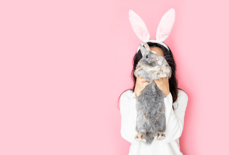 funny happy girl with bunny ears holding gray rabbit on pink background.の写真素材