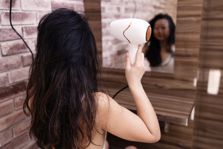 Back view of asian woman blow dry hair by hair dryer and looking in front of the mirror in the bathroomの写真素材