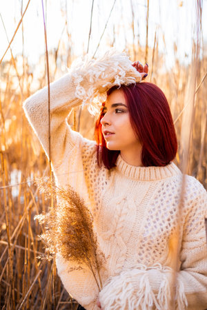 Elegant young woman in a white woolen sweater poses in the autumn park on the background of reeds. Autumn fashion and beauty.の写真素材