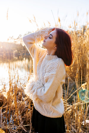 Elegant young woman in a white woolen sweater poses in the autumn park on the background of reeds. Autumn fashion and beauty.の写真素材