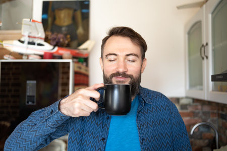 Photo of handsome positive optimistic nice glad attractive guy holding cup of fresh coffee in hands looking in window standing in kitchenの写真素材