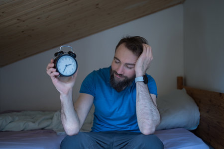 Sleepless and desperate young caucasian man stays awake at night, unable to sleep, feeling frustrated and anxious, looking at clock, insomniac in stress and sleep disorder concept.の写真素材