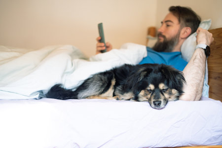 Man in a blue shirt lying on a bed with a dog and using a smartphone.の写真素材