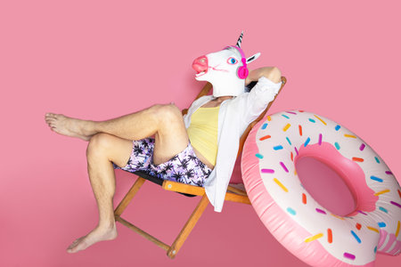 Young funny tourist man in unicorn head mask with headphones lie on deckchair isolated on pink background studio portrait. Summer vacation sea rest sun tan conceptの写真素材