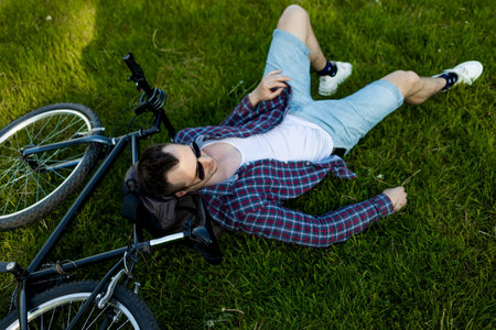 Young man lying on lawn next to bike in summer park. happy person resting after cycling, eyes...