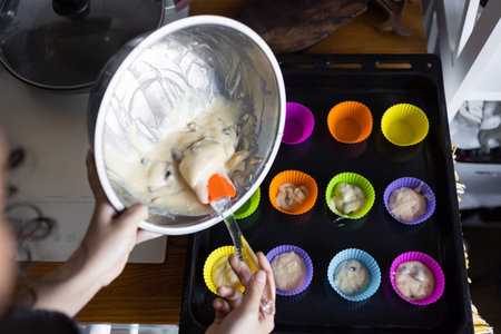 making of delicious cupcake in kitchen. Muffin tin with liner, ingredients, and utensils are seen. A woman is adding mixture into the holesの写真素材