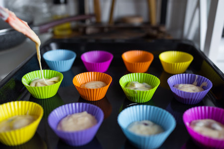 making of delicious cupcake in kitchen. Muffin tin with liner, ingredients, and utensils are seen. A woman is adding mixture into the holesの写真素材