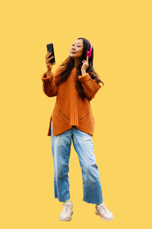 Focused young asian female in sweater wear listening to music in headphones and browsing mobile...