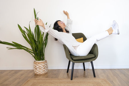 Attractive Asian woman sitting on modern chair and hands up raised arms from happiness in living room, Excited woman winner success concept.の写真素材