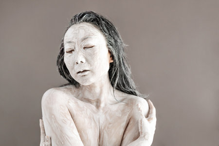 Art portrait of asian woman covered in clay isolated over grey background. Woman face like cracked pottery cup holding by hand.の写真素材