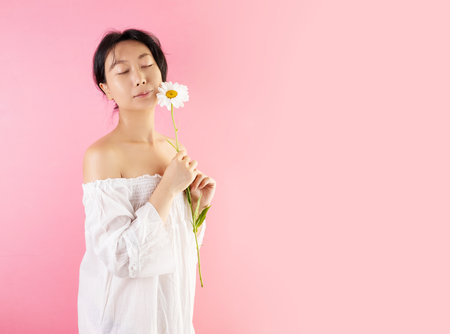 Closeup of happy cheerful pretty young asian woman posing on pink background, touching face with white daisy flower and smiling.の写真素材