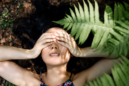 Young asian woman covering her eyes with her hands while lying in the forest
