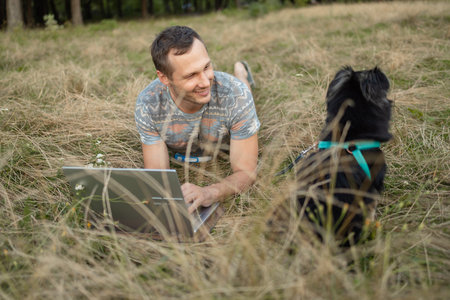 Young man with dog working outdoors with laptop in park.