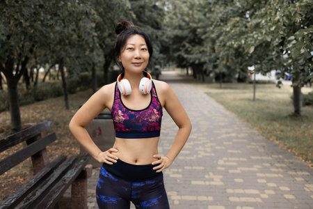 Portrait of a young beautiful asian woman in sportswear standing on a bench in the parkの写真素材