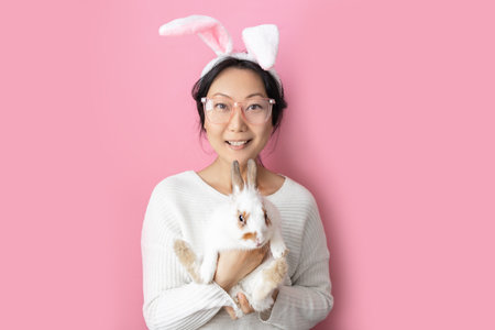 Asian woman wearing bunny ears and holding white rabbit on pink background.の写真素材